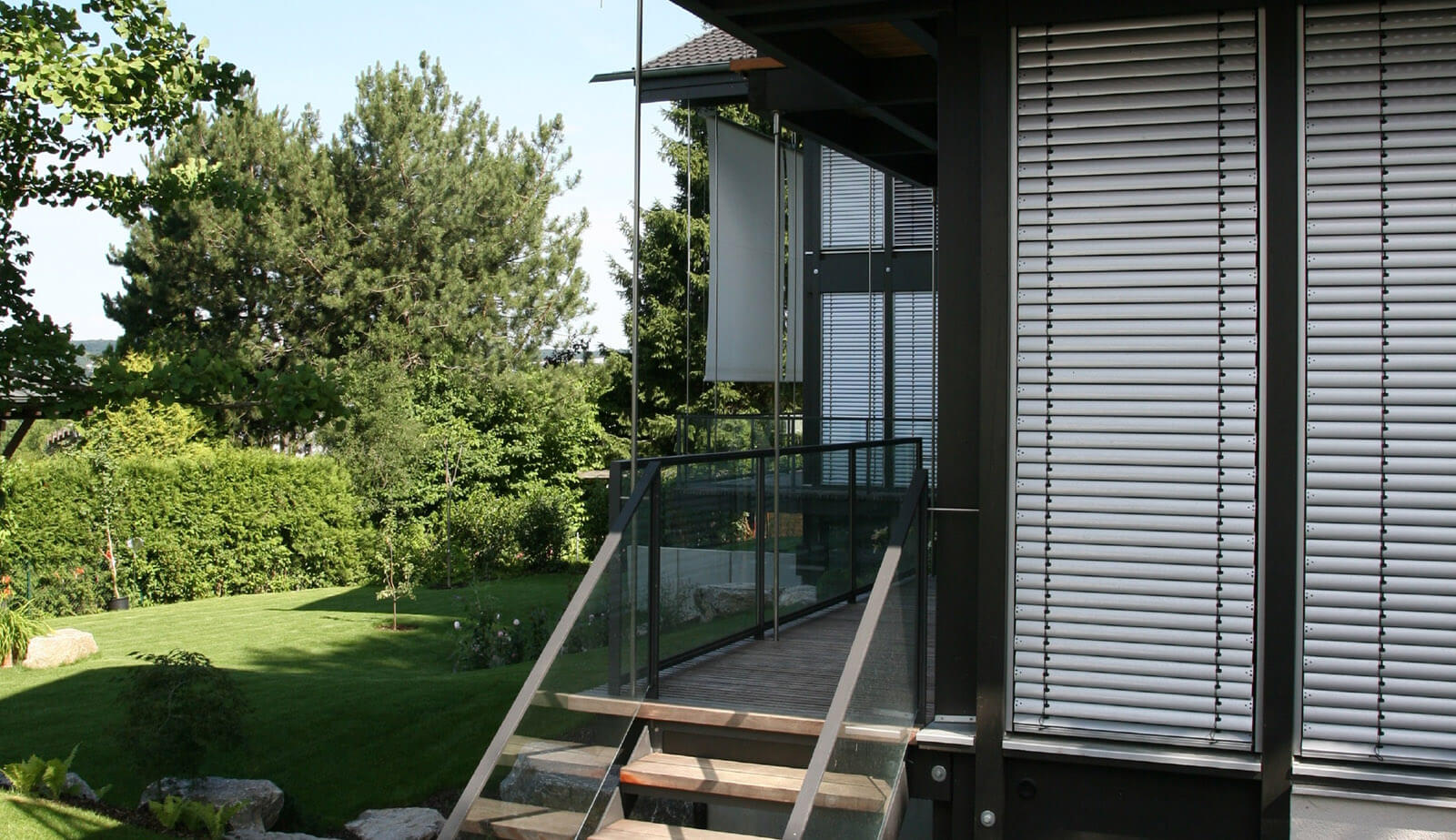 Doppelvilla: Außentreppe zum Holzsteg mit Glasgeländer neben Aluminiumjalousien