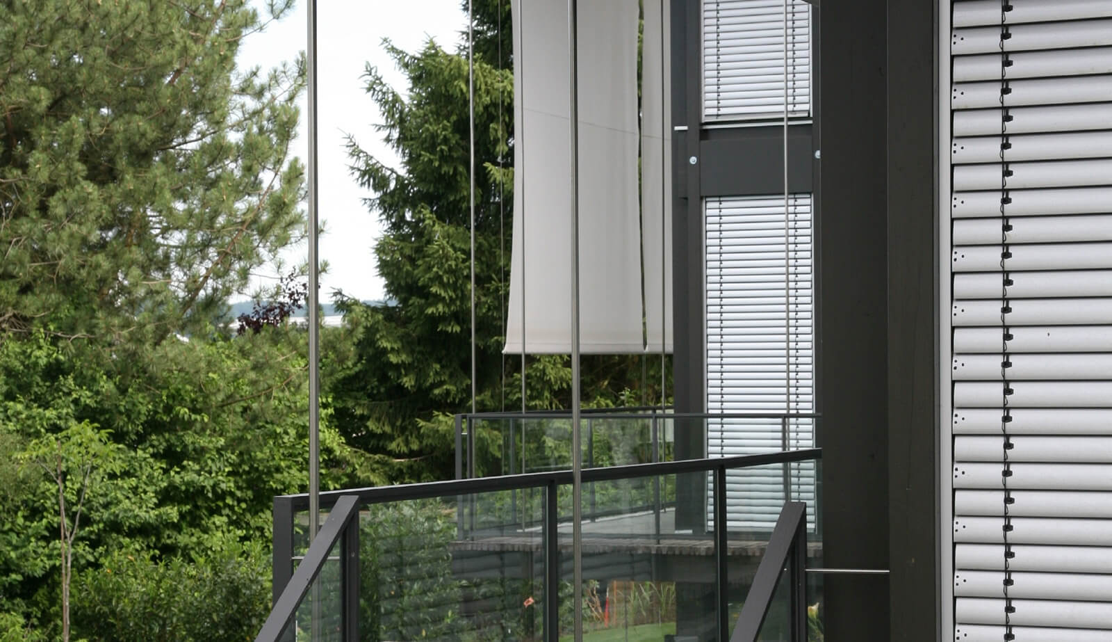 Doppelvilla: Glasgeländer, Sonnensegel und Jalousien vor Nadelbäumen
