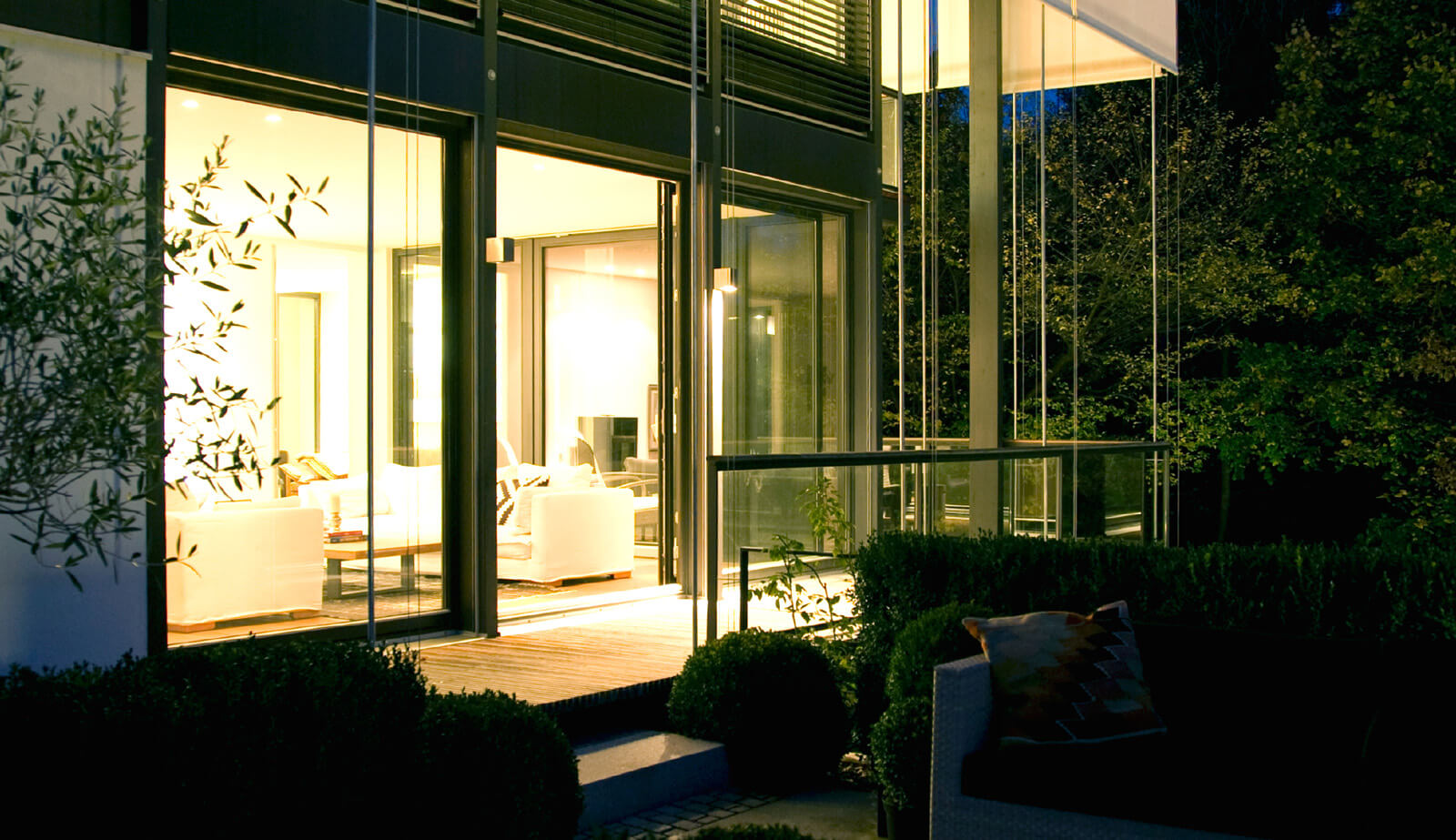 Flachdach-Kubus: erleuchtete Glasfront hinter Garten bei Nacht