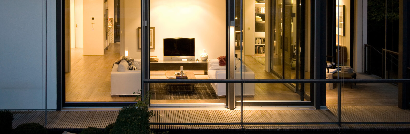 Flachdach-Kubus: beleuchteter Wohnraum hinter Glasfront mit Holzterrasse am Abend