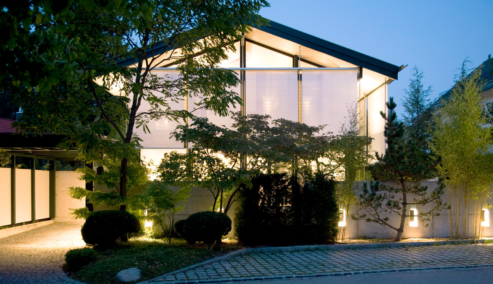 On a small plot: saddle roof house with glazed gable and lit front garden at dusk