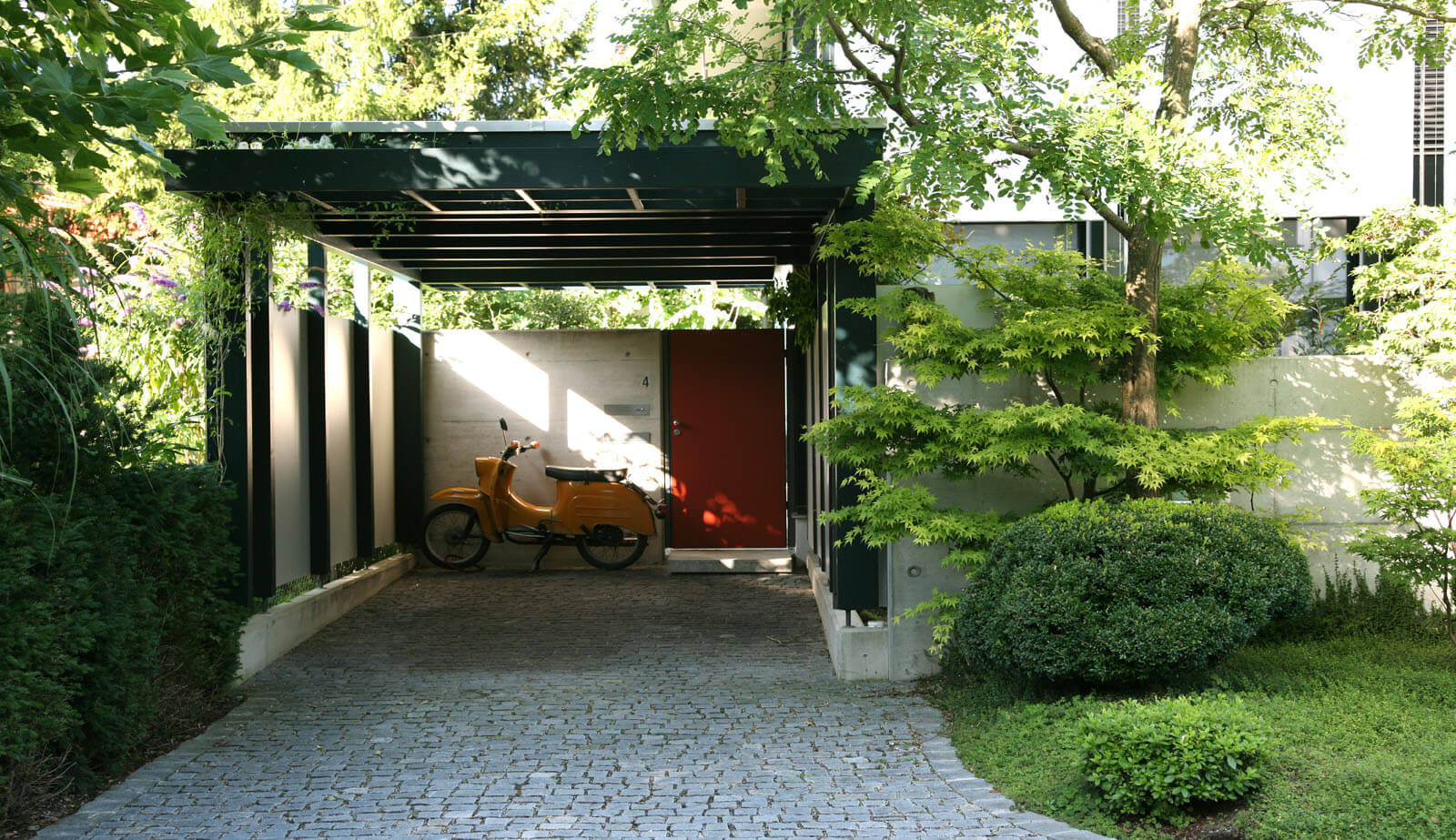 On a small plot: carport with pergola, red entrance door and cobbled driveway