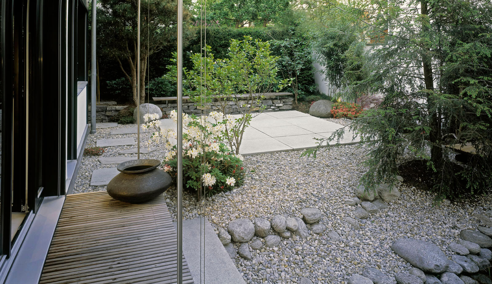 On a small plot: gravel garden with boulders and timber walkway along the glass facade