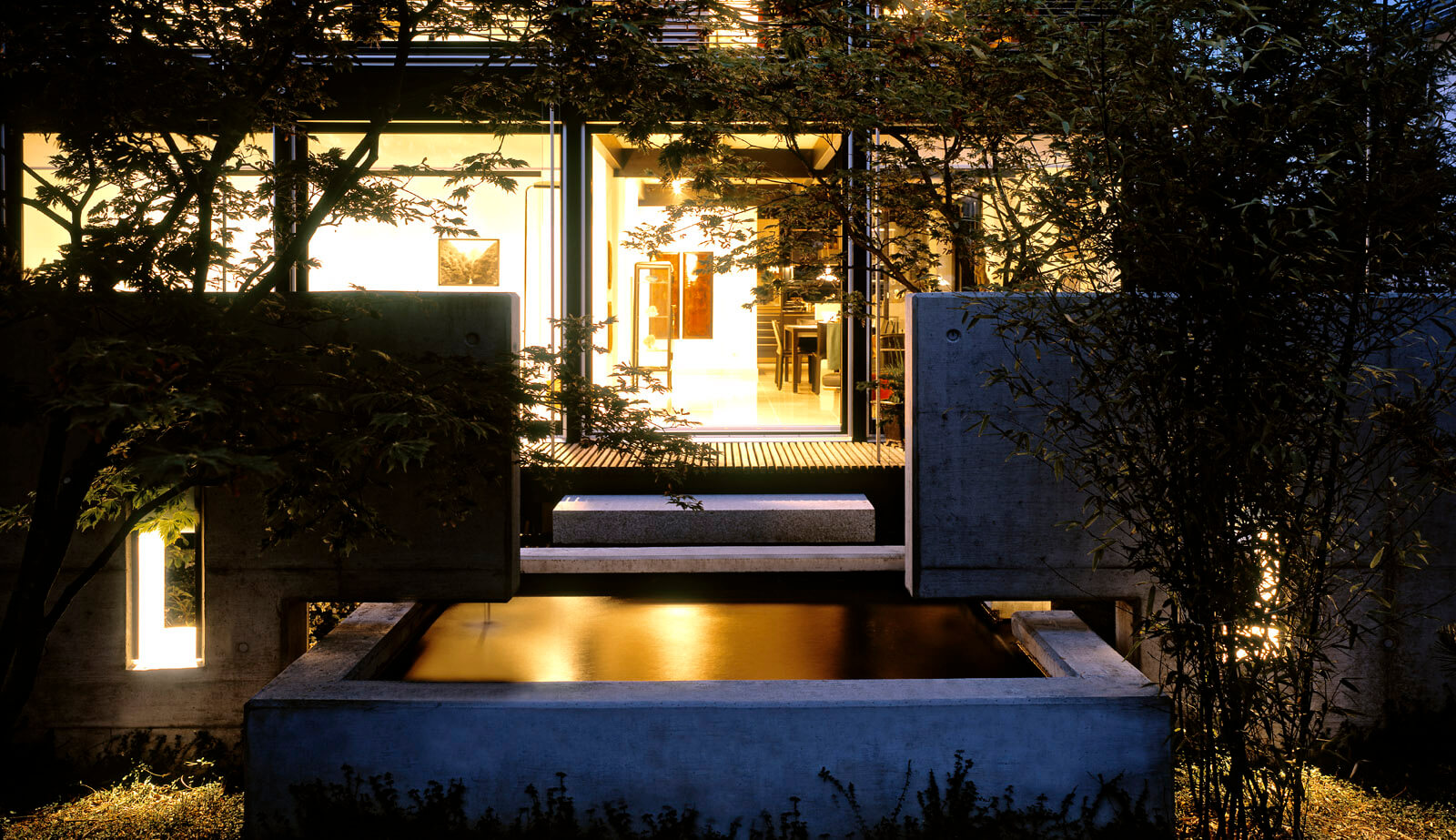 On a small plot: concrete fountain and steps in front of lit living room at night