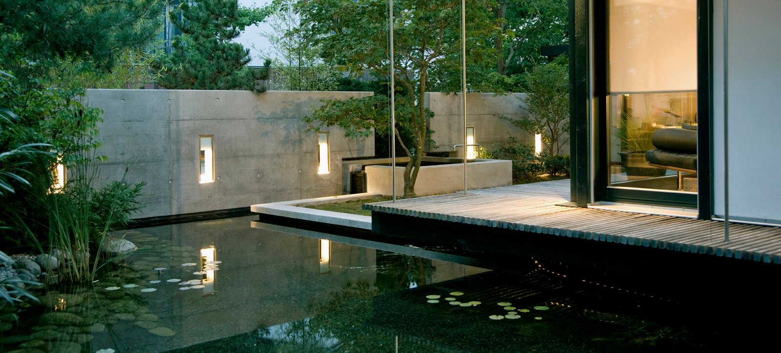 On a small plot: pond and timber deck in front of concrete wall with wall lights