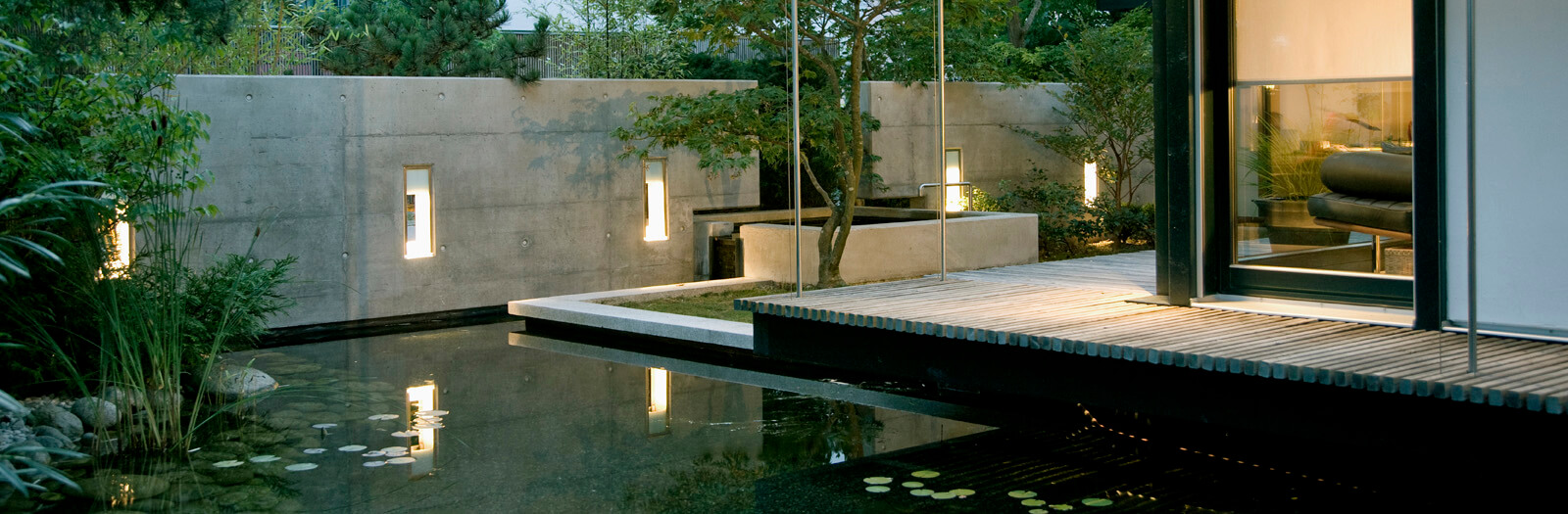 On a small plot: lit pond with timber deck in front of the glass facade at dusk