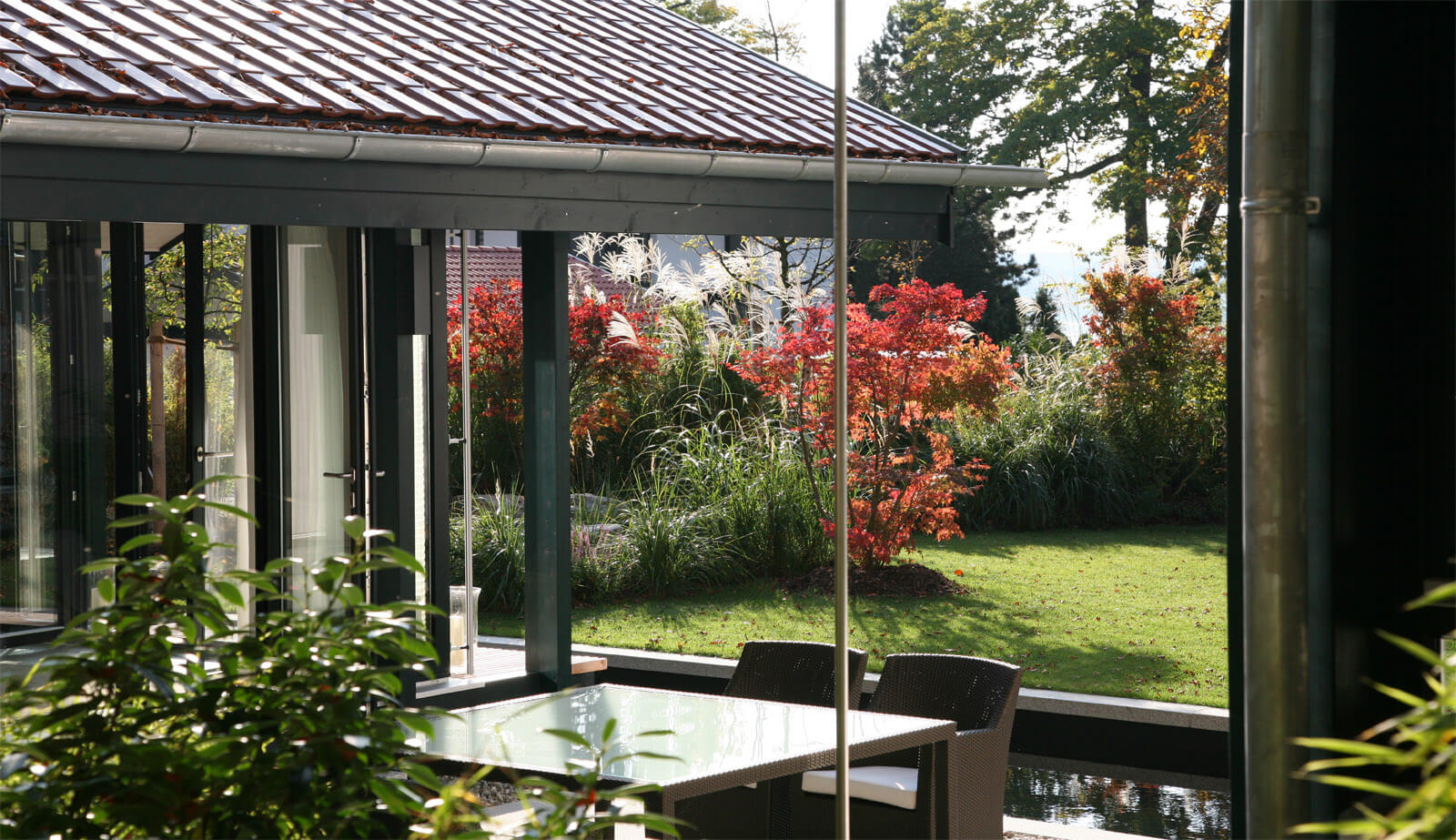 Haus mit Gästehaus: Terrasse mit Glastisch unter Ziegeldach, Herbstgarten dahinter
