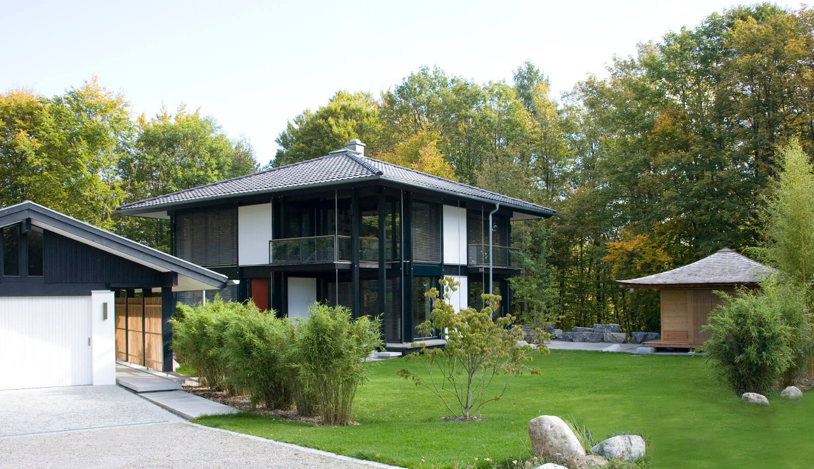 Zeltdachvilla: Haus mit Garage, Gartenhaus und Rasen vor herbstlichem Wald